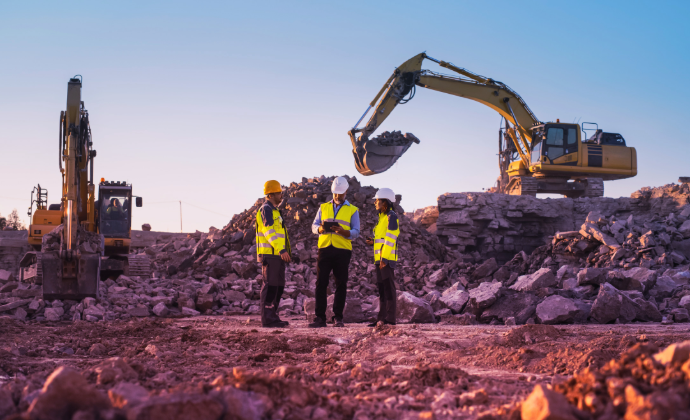 Three construction workers in high-visibility vests and hard hats stand on a rubble-covered site, discussing plans as one excavator moves rocks and another remains stationary under a clear, gradient sky