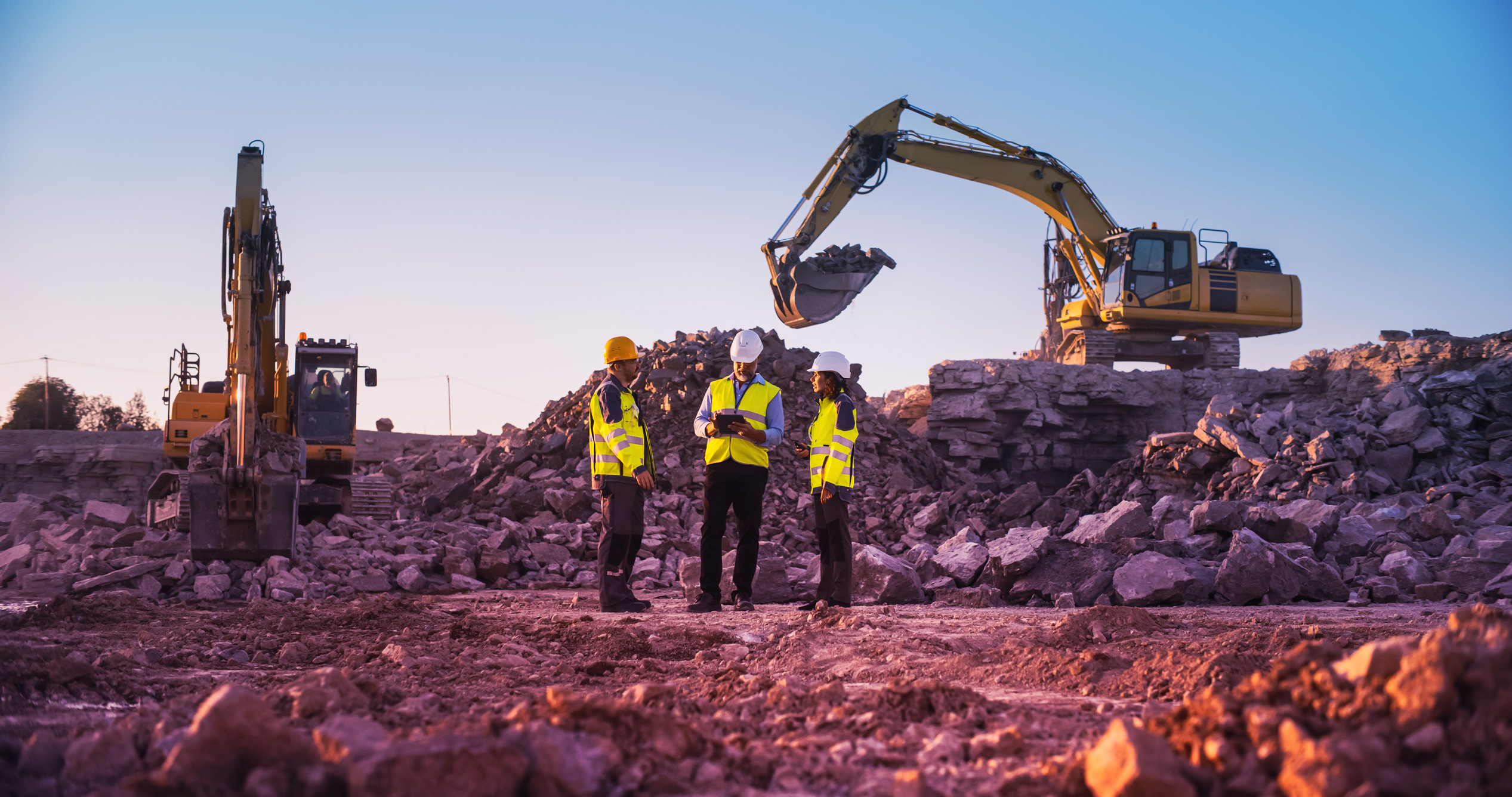 Three construction workers in high-visibility vests and hard hats stand on a rubble-covered site, discussing plans as one excavator moves rocks and another remains stationary under a clear, gradient sky